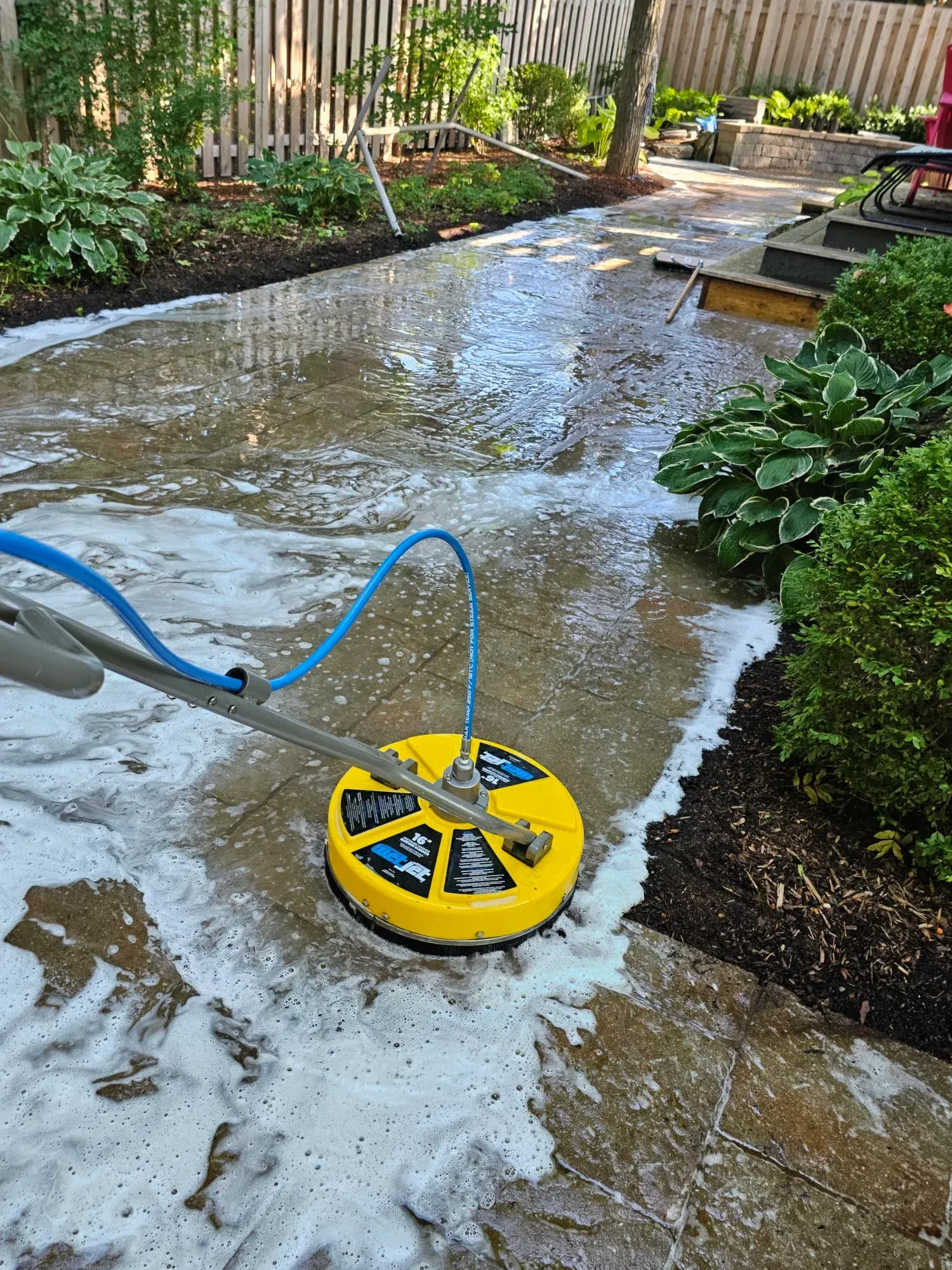 Rotary surface cleaner working across a foamed paver walkway, clean wet stone emerging behind the tool